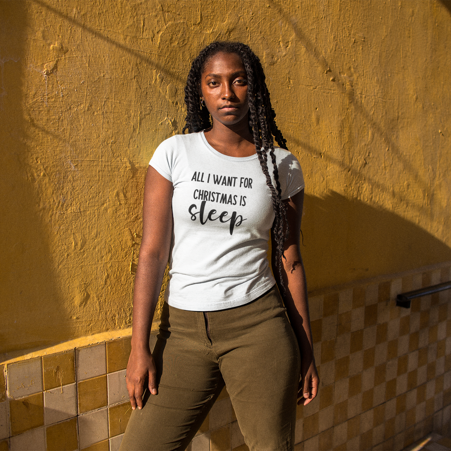 Black woman with braids in front of yellow wall, wearing white shirt with 'All I want for Christmas is sleep' print by KMLeon.