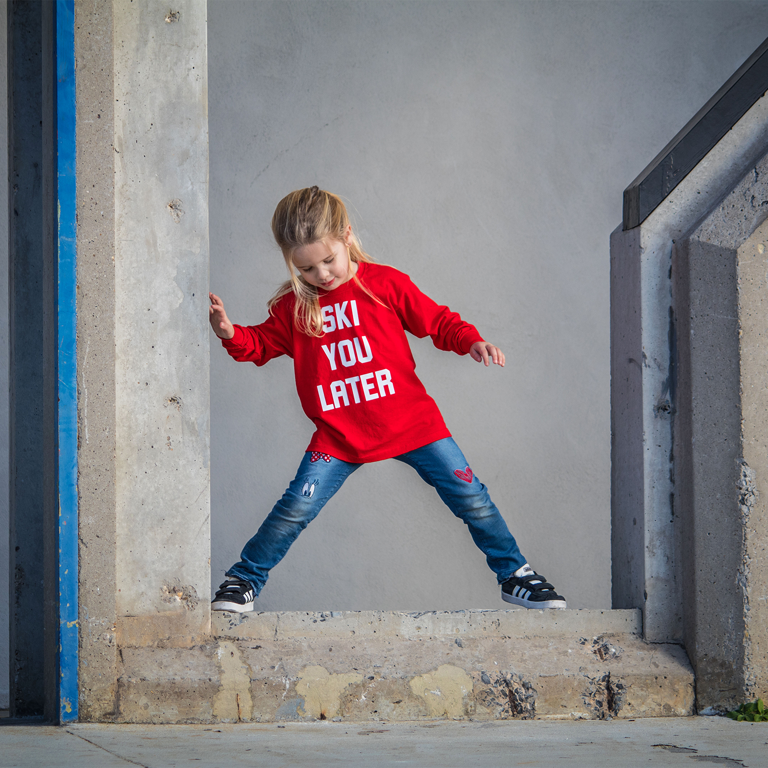 Blonde girl with red shirt with 'Ski you later' print by KMLeon standing on concrete.