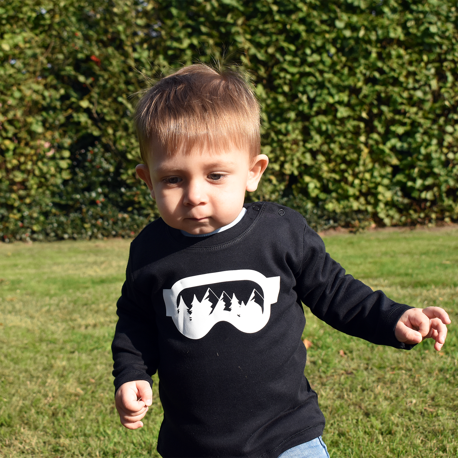 Little boy wearing black shirt with longs sleeves withh ski goggles print by KMLeon walking in park.