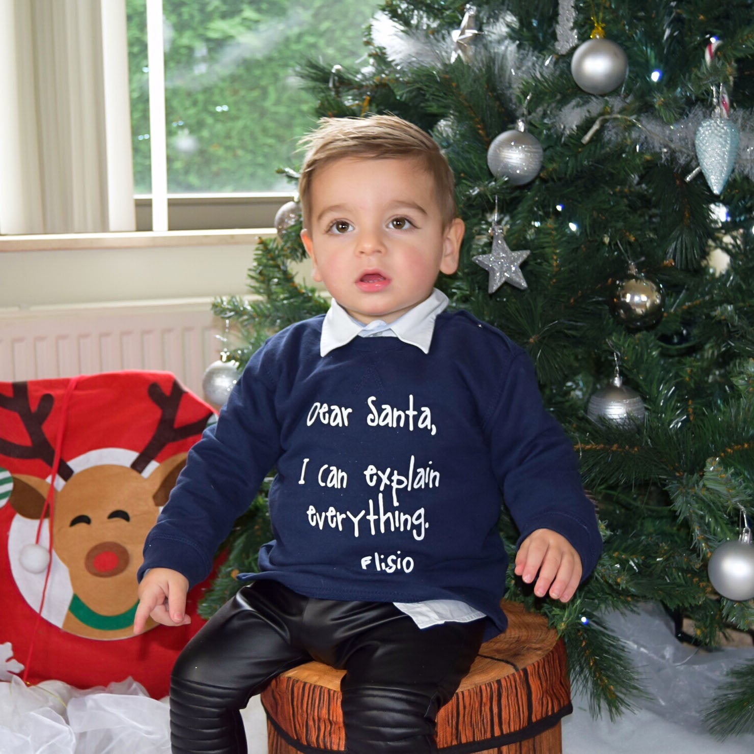 Boy with Navy sweater with 'Santa, I can explain everything' print by KMLeon in front of christmas tree.