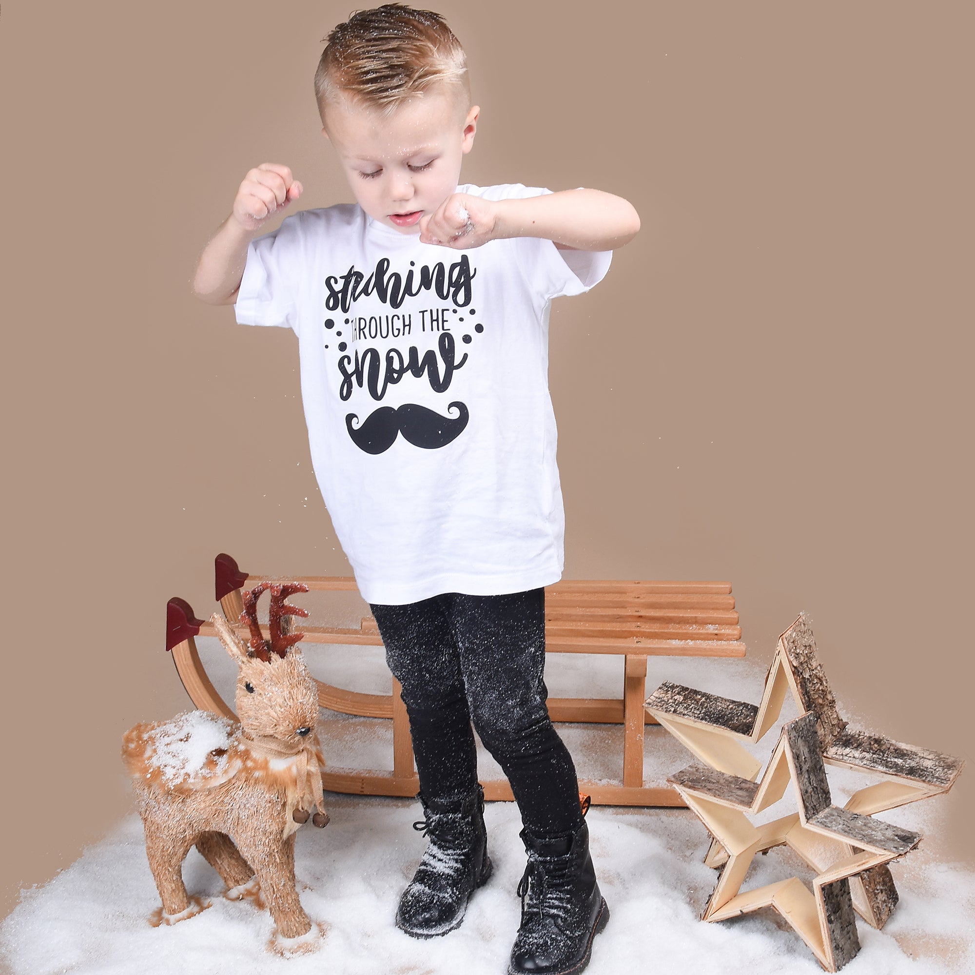 Blonde boy playing with snow, wearing white shirt with 'Staching through the snow' print by KMLeon.